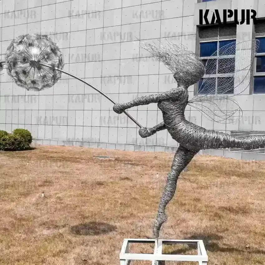 Art Deco stainless steel fairy dandelion sculpture standing on a paved outdoor plaza with reflective surface catching sunlight