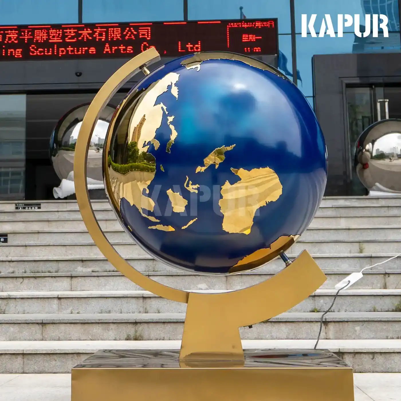 Mirror-polished stainless steel rotating globe standing on a concrete base in an outdoor plaza, showing the reflective surface and rotating mechanism