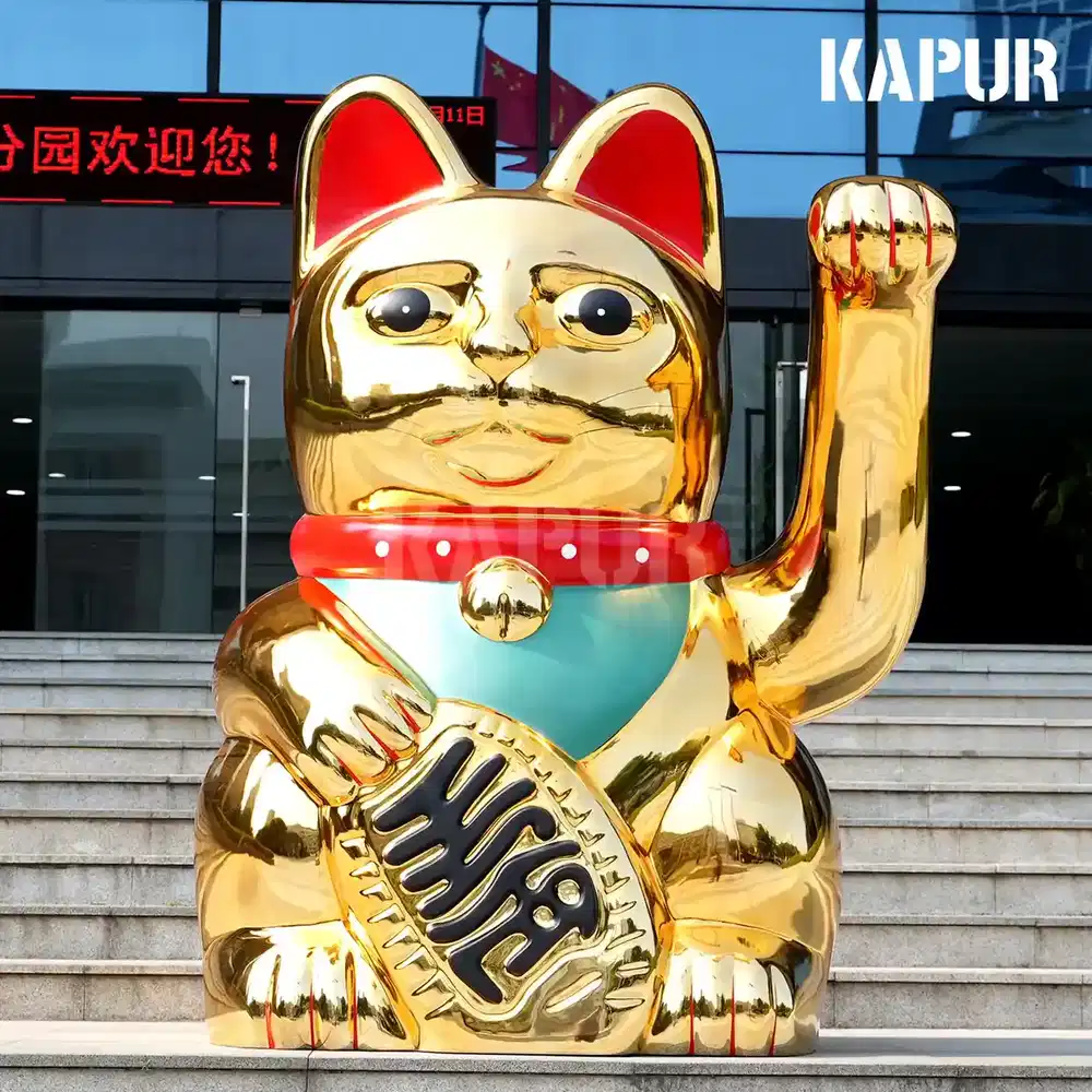 Polished stainless steel beckoning cat sculpture standing on a paved outdoor plaza, facing forward with one hand raised