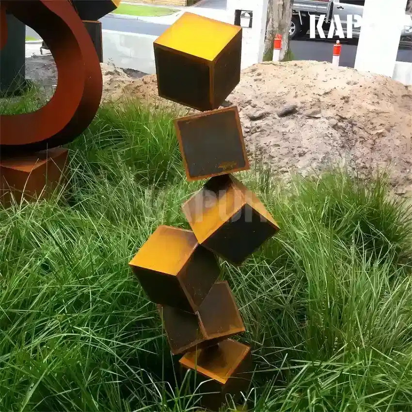 Large corten steel sculpture in the form of stacked building blocks, standing in an outdoor garden with natural vegetation