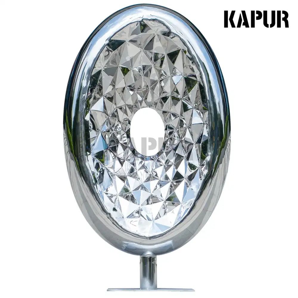 Polished stainless steel oval gemstone sculpture standing on a paved outdoor plaza, reflecting light and surrounding architecture