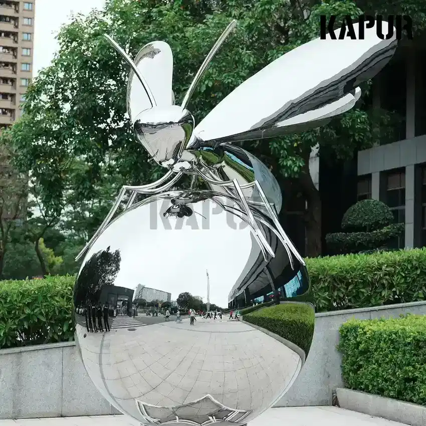 Mirror-polished stainless steel bee sculpture in a landscaped plaza