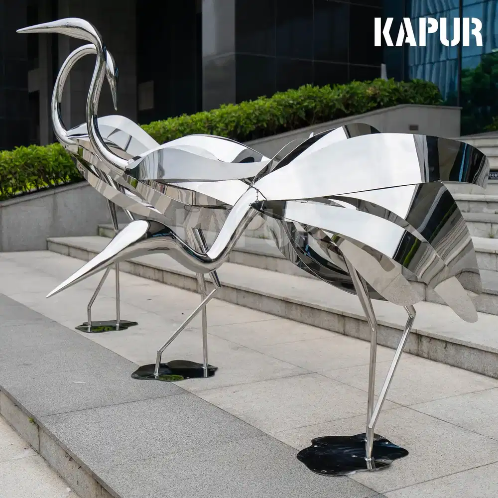 Polished stainless steel ostrich animal sculpture standing in a modern garden setting with reflective surface catching sunlight
