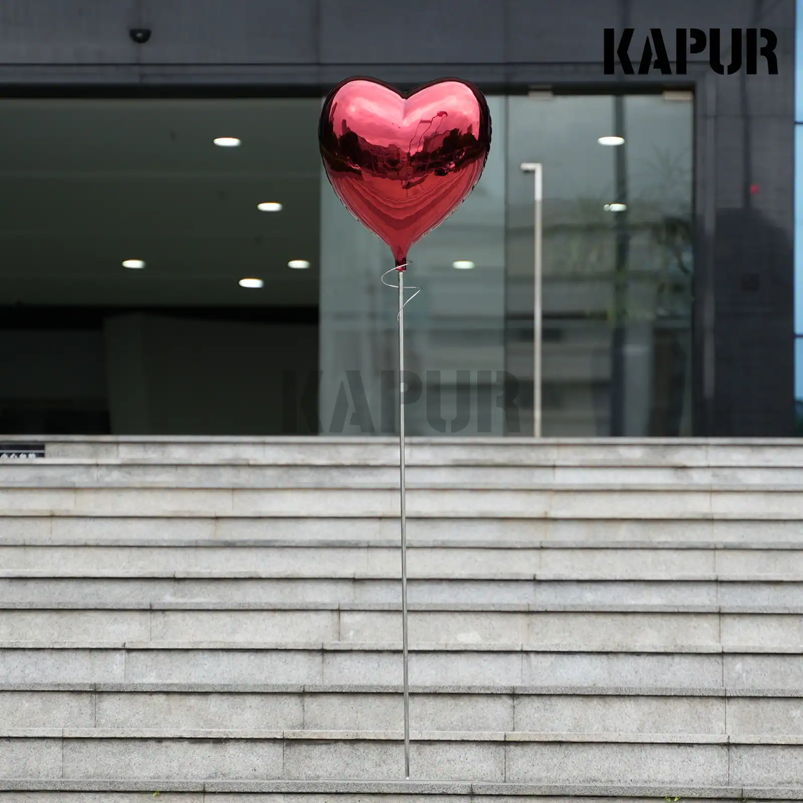 Polished stainless steel love balloon sculpture standing on a paved outdoor plaza, reflecting the sky and surrounding architecture