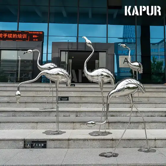 Polished stainless steel flamingo animal sculpture standing in a modern garden setting with reflective surface catching sunlight