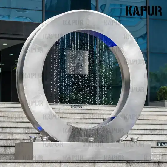 Round stainless steel water curtain sculpture with flowing water effect, standing in a modern outdoor plaza