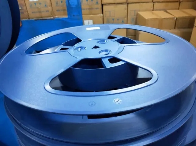 Large blue plastic reel with cutout spokes in a warehouse, stacked cardboard boxes visible