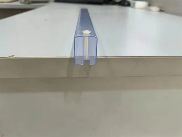 Translucent plastic U-channel profile strip resting on the edge of a white table, with a small white plug fitting visible