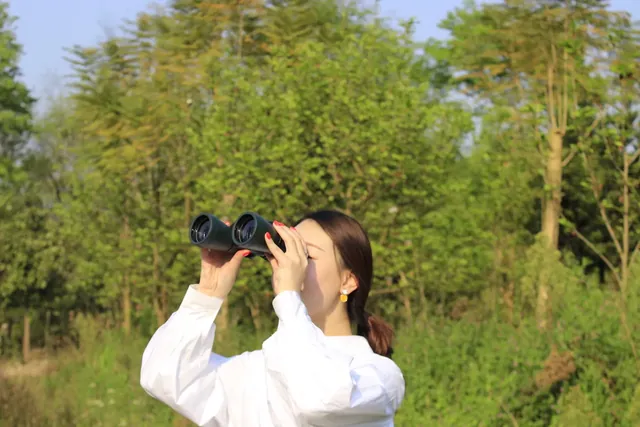 Woman looking through black binoculars against a background of green trees