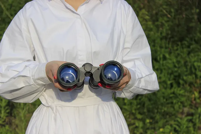 A person wearing a white shirt holds a pair of black binoculars with blue-tinted lenses outdoors
