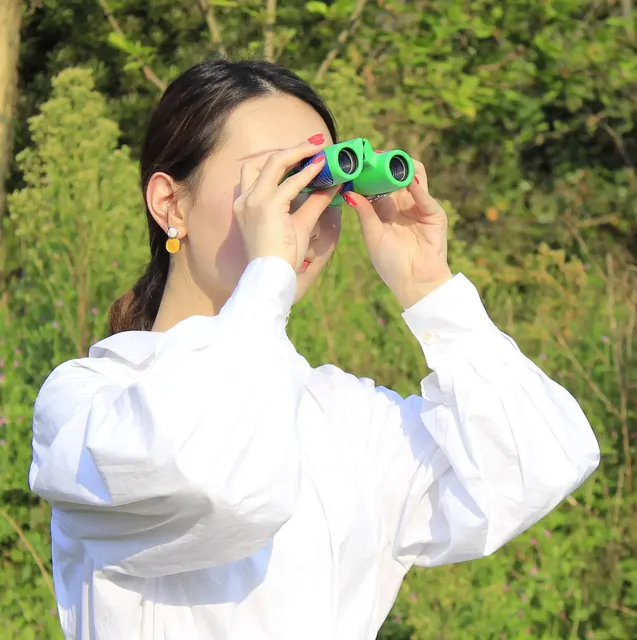 Woman in a white shirt looking through green binoculars in a forest