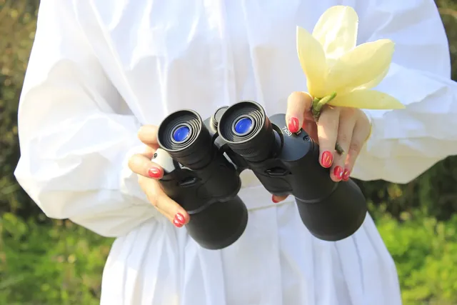 Person in white shirt holding black binoculars and a yellow flower outdoors