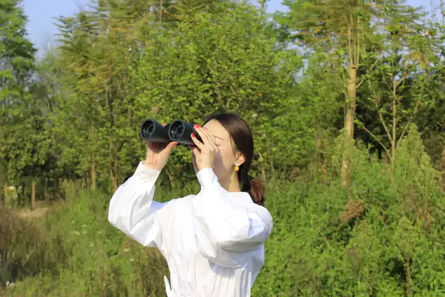 Woman in white shirt looking through binoculars in a grassy field with trees