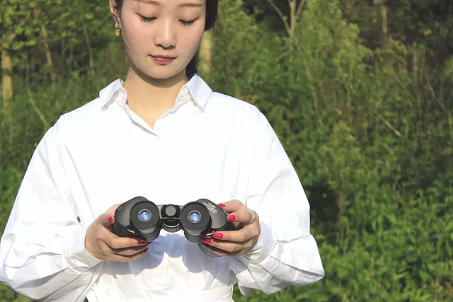 Woman in white shirt holding a pair of black binoculars outdoors against a green background