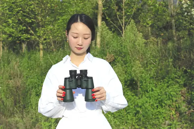 Woman in white shirt holding green binoculars outdoors against a background of trees and bushes