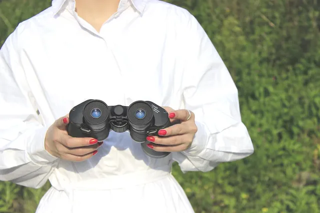 Person in a white shirt holding black binoculars outdoors against a grassy background