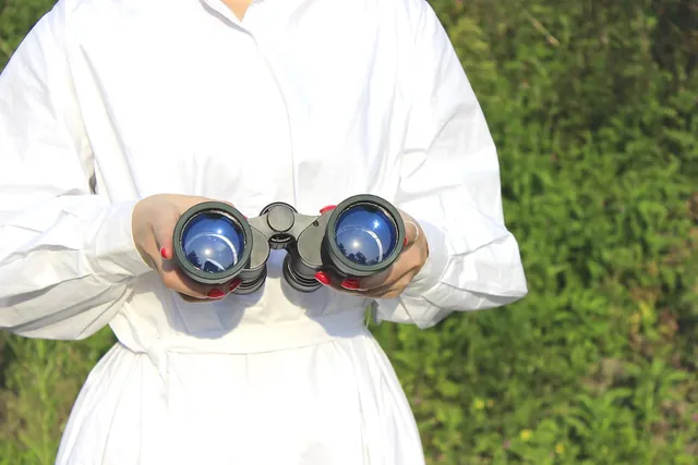 Person in white shirt holding binoculars outdoors with green foliage in background