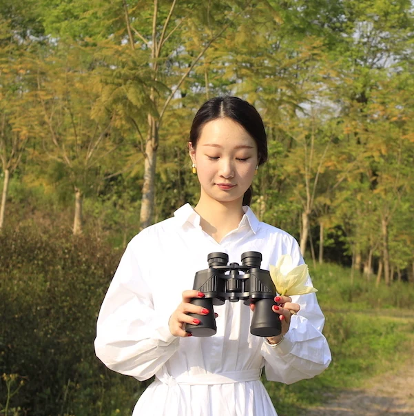 Woman in a white shirt holding black binoculars outdoors with a background of trees
