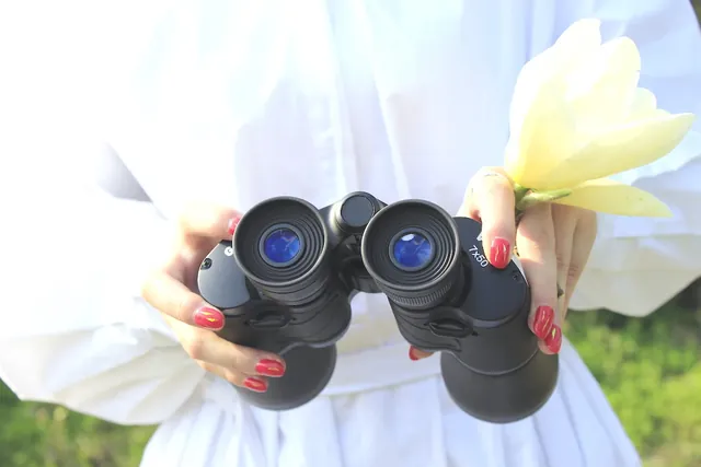 Black binoculars held by a person with red nail polish, alongside a pale yellow flower