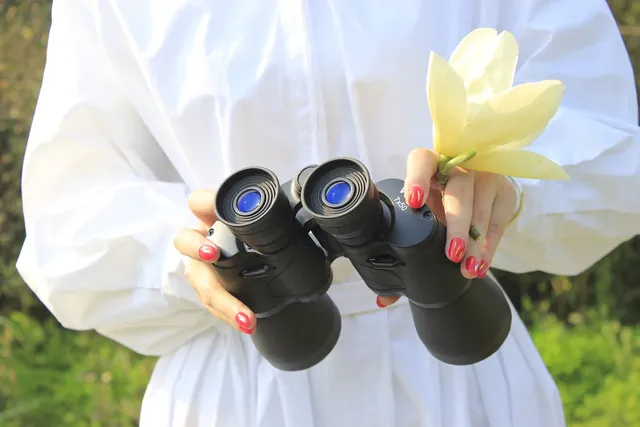A person wearing white clothing holding a pair of black binoculars and a large yellow flower