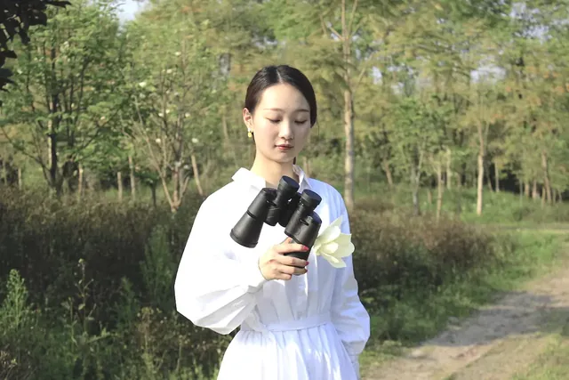 A woman in a white dress stands outdoors holding a pair of black binoculars and a white flower