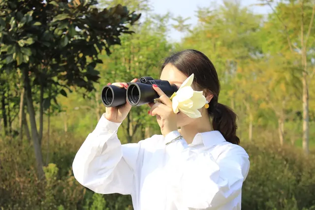 Woman in a white shirt looking through binoculars while holding a yellow flower outdoors