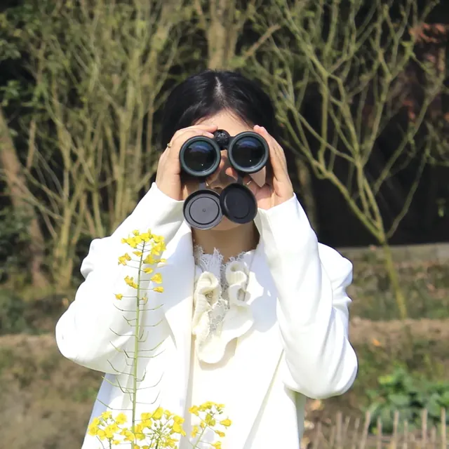 A woman in a white jacket holding black binoculars to her eyes outdoors