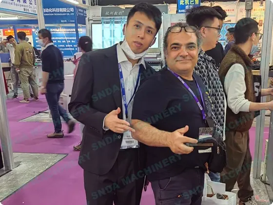 Two men posing together at a trade show exhibition with booths and other visitors in the background