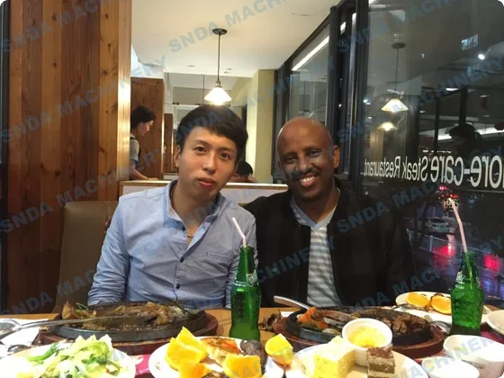 Two men seated at a restaurant table with sizzling plates, salad, orange slices and green glass bottles