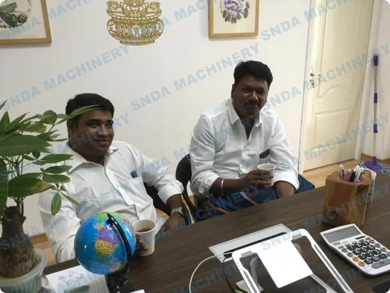 Two men in white shirts seated at a cluttered office desk with a globe, calculator and coffee mug