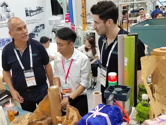 Three men wearing lanyards stand behind a table of industrial supplies at a busy trade show booth