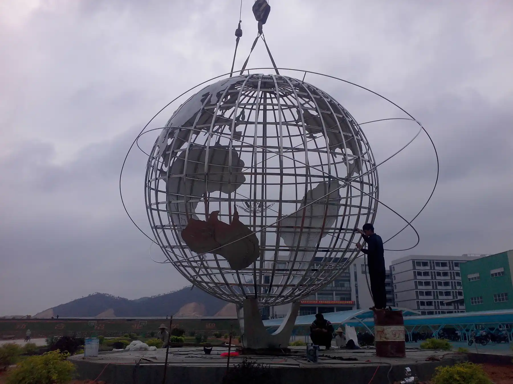 A large metal globe sculpture being lifted by a crane outdoors, with a building and mountains in the background.