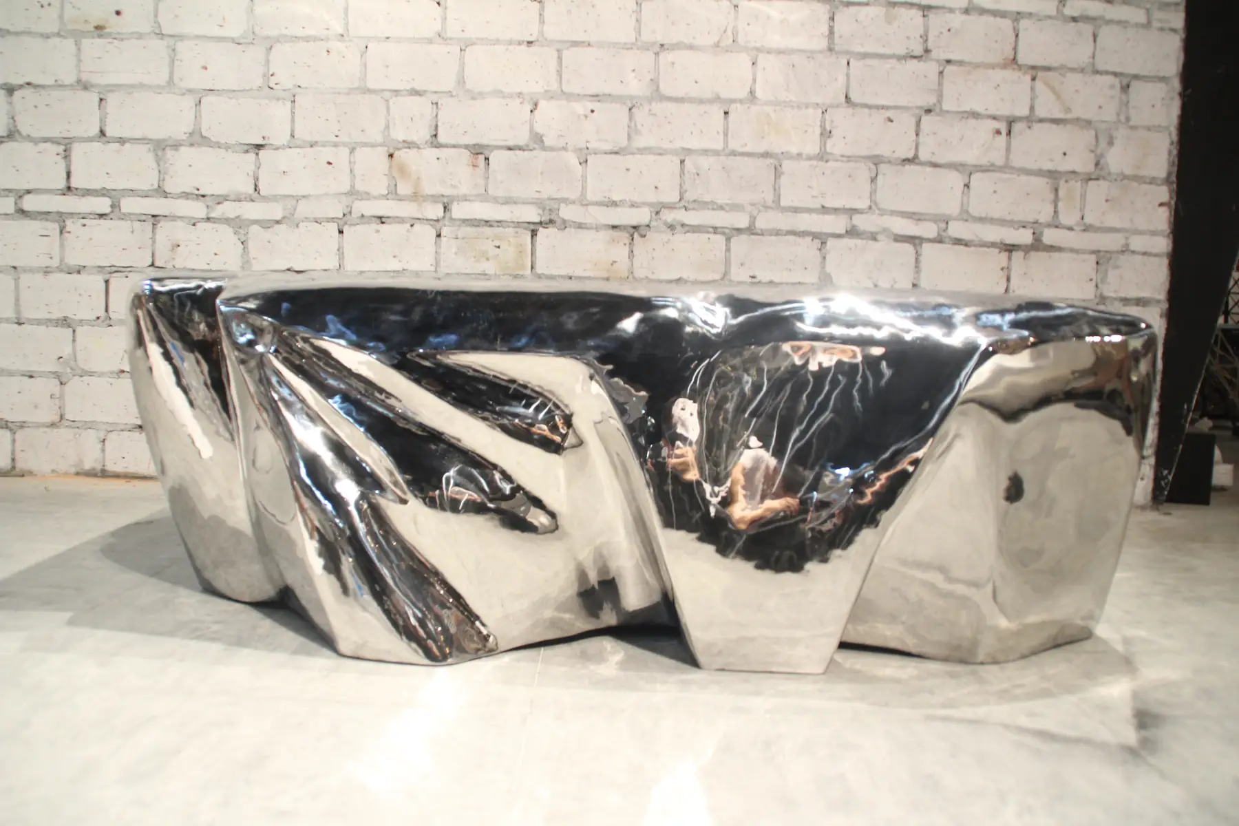 A shiny, abstract metal sculpture with a reflective surface, set against a white brick wall.
