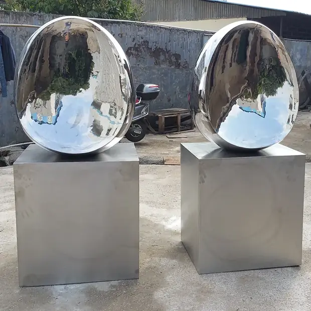 Two large, shiny, egg-shaped metal sculptures on square metal pedestals outdoors.