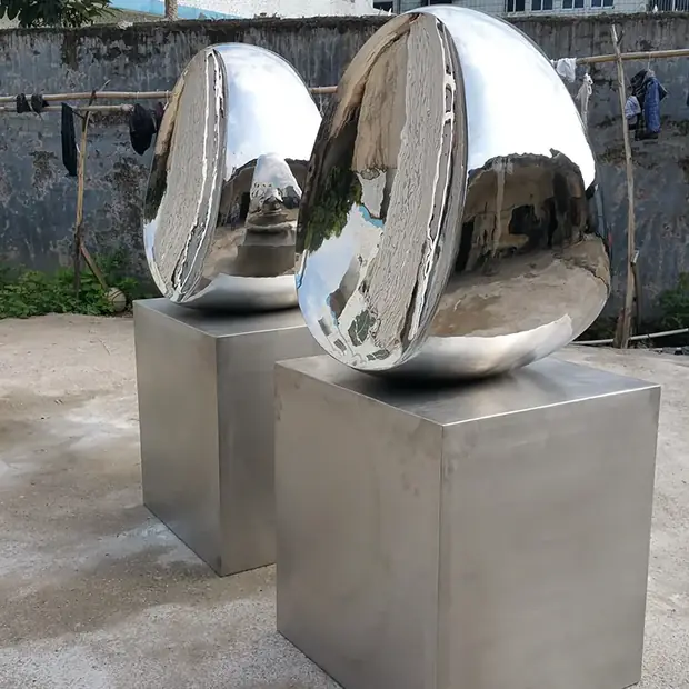 Two large, reflective, egg-shaped metal sculptures on square pedestals outdoors.