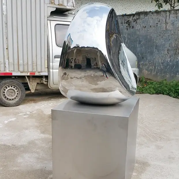 A large, shiny, abstract metal sculpture sits on a metal pedestal outdoors, with a truck and wall in the background.