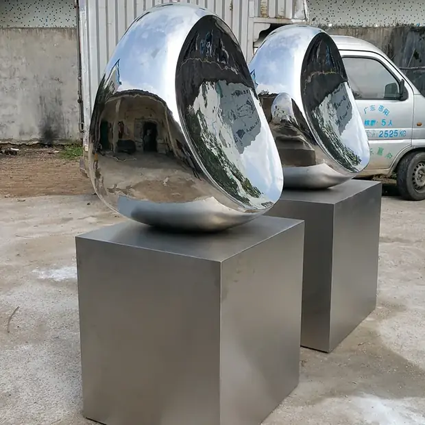 Two large, shiny, egg-shaped metal sculptures on gray pedestals outdoors.