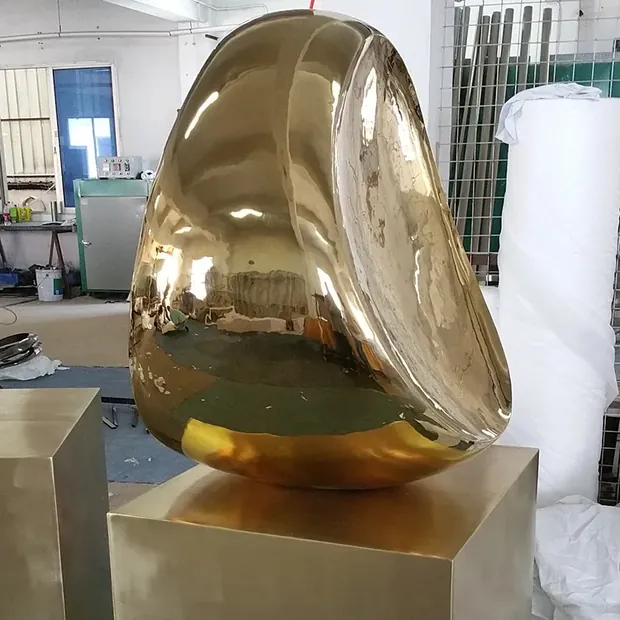 Large golden reflective sculpture on a pedestal in a workshop.