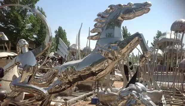 Large stainless steel horse-drawn carriage sculpture on a plaza with people walking nearby