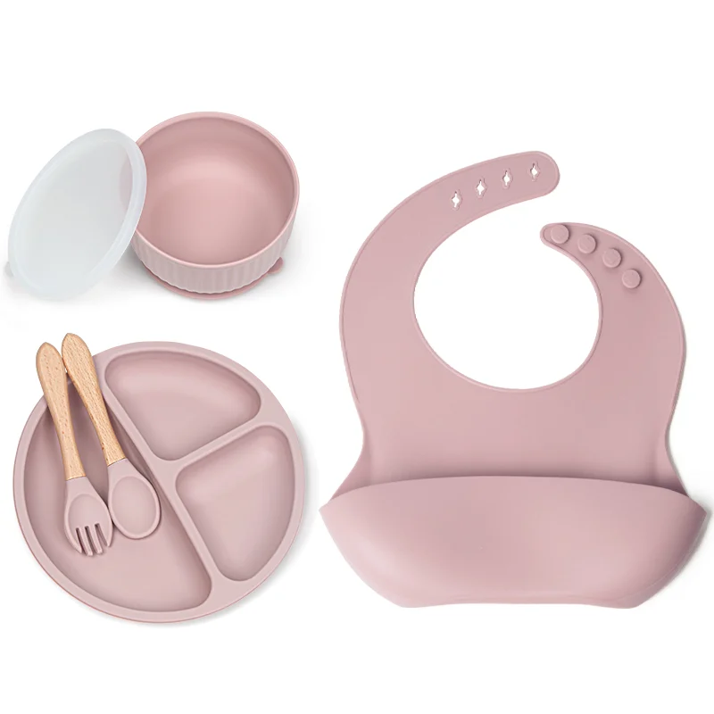 Silicone baby feeding set with bowl, lid, bib, spoon and fork laid out on a white surface