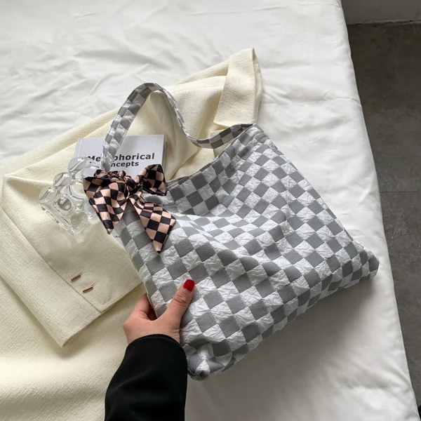 Hand holding a gray and white checkerboard canvas shoulder bag with a brown bow charm on a bed.