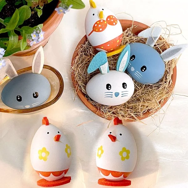 Decorated Easter eggs styled as bunny and chick characters in a straw-filled terracotta bowl with spring flowers