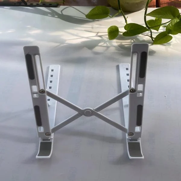 White folding X-frame adjustable laptop stand photographed upright on a table with green plant leaves in the background