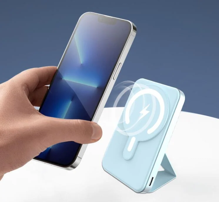 A hand holding a smartphone next to a light blue magnetic wireless power bank with a kickstand and charging graphic.