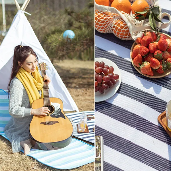 Woman playing guitar on a striped picnic blanket outdoors next to a tent and fruit.