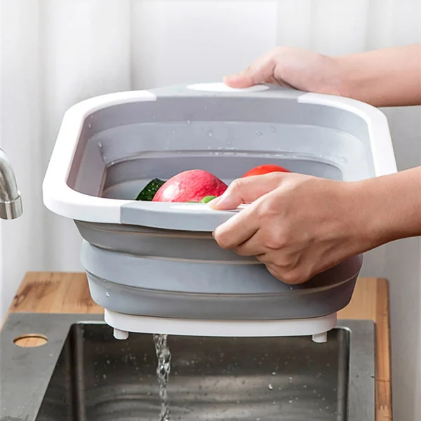 Hands holding a gray collapsible drain basin filled with fresh fruit over a stainless steel kitchen sink