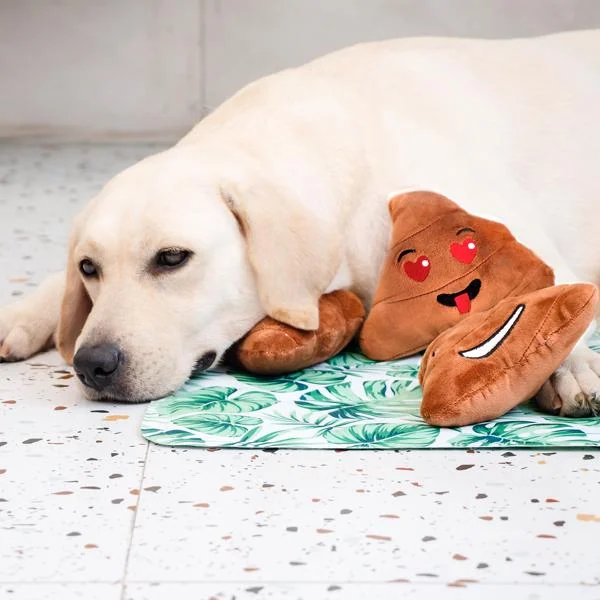 A yellow Labrador lying on a green patterned mat, resting its head near a brown plush poop dog toy with heart eyes.