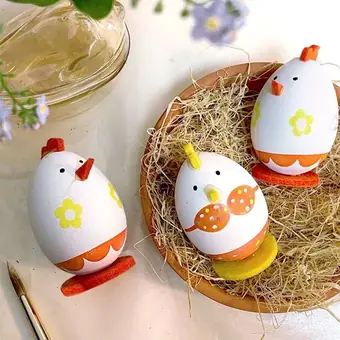 Three white Easter eggs decorated as chicks with orange and yellow details, one in a terracotta bowl with straw