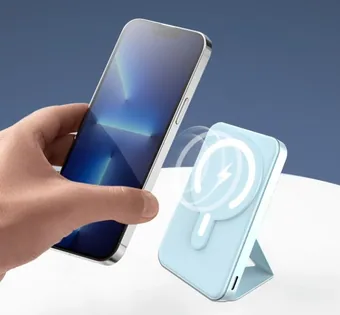 A hand holding a smartphone next to a light blue magnetic wireless power bank with a kickstand and charging graphic.