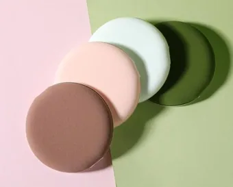 Four round makeup sponge powder puffs in brown, pink, mint and dark green arranged diagonally on a pink and sage background